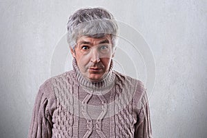 A horizontal portrait of senior handsome man standing against white wall wearing sweater looking with astonishment into camera. Pe