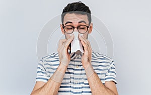Horizontal portrait of unhealthy handsome man wearing striped shirt and glasses, blowing nose into tissue. Male have flu, virus or