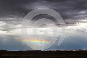 Horizontal rainbow across the middle of a rain squall near the horizon in desert with distant cliffs lit up and foreground dark - photo