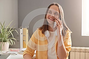 Horizontal shot of adorable winsome female with charming smile, resting at work, talking on smart phone, looking aside with