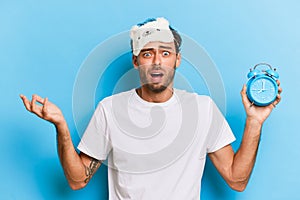 Horizontal shot of amazed caucasian young bearded man in sleeping mask holds alarm clock shows time looks frustrated and