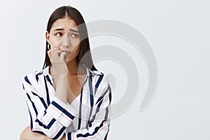 Horizontal shot of anxious good-looking female student in striped blouse, biting fingernail nervously, frowning and