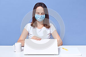 Horizontal shot of attentive freelancer closing her laptop, sitting at table, having cup nearby, wearing antibacterial mask, being