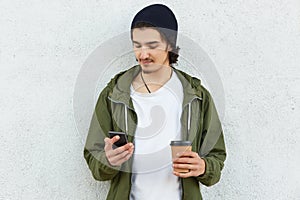 Horizontal shot of attractive younsgter in stylish headgear and green jacket, holds modern smart phone in one hand and takeaway co