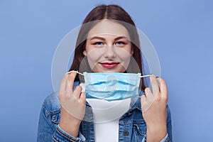 Horizontal shot of beautiful woman puts on medical mask isolated over blue background, girl trying to avoid flu or coronavirus.