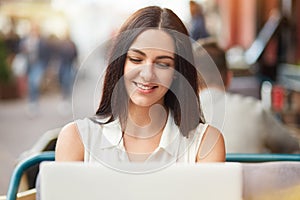 Horizontal shot of beautiful young Caucasian female sits in front of opened laptop computer, smiles gently, messages in social net