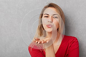 Horizontal shot of beautiful young woman with appealing appearance, makes air kiss, blows at camera, wears red sweater, o