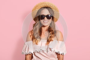 Horizontal shot of beautiful young woman in summer shirt with bare shoulders and sun hat, looking smiling at camera, has happy