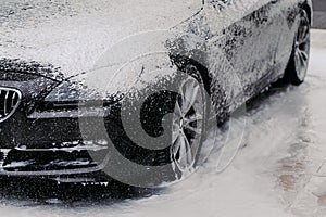 Horizontal shot of black automobile covered with bubble soap foam at carwash. Detail cleaning. Purifying concept