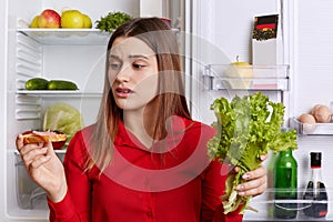 Horizontal shot of brunette female with upset expression looks at sandwhich, keeps lettuce, wants to eat meat, keeps to diet, does