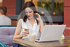 Horizontal shot of busy female freelancer holds mobile phone, text messages online, uses laptop computer, drinks milkshake, poses