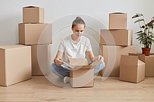 Horizontal shot of Caucasian woman with bun hairstyle wearing white T-shirt sitting on the floor near cardboard boxes with stuff,