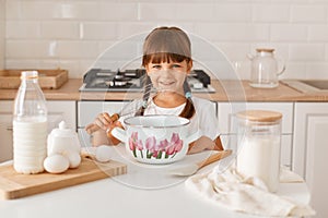 Horizontal shot of charming little girl preparing dough for baking, posing with corolla in hands, mixing ingredients for cake,