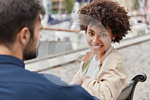 Horizontal shot of cheerful Afro American girl smiles broadly, sits opposite Caucasian unshaven guy, pose at bench