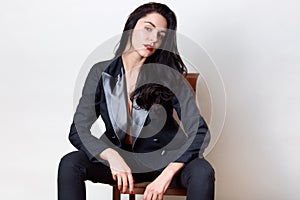 Horizontal shot of confident brunette girl with long beautiful hair, winsome woman sitting on chair and looking directly at camera