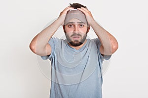 Horizontal shot of confused helpless young man being in panic, holding arms on head, looking directly at camera, wearing sleeping