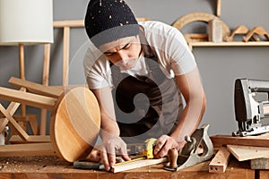 Horizontal shot of creative young carpenter wearing black cap, apron and white shirt working in workshop, measuring wooden plank