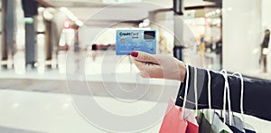 Horizontal shot of crop female hand holding face side of readable credit card at mall background. Female shopaholic using bank