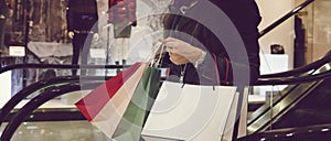 Horizontal shot of crop woman in leather black jacket holding many shopping bags and moving down on escalator in mall. Concept of