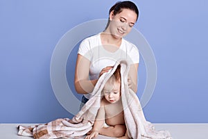 Horizontal shot of cute happy baby kid sitting wrapped in white and brown towel after bath, mum wiping and drying kid isolated