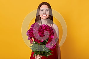 Horizontal shot of cute young woman with flowers standing isolated over yellow background, female giving bouquet of maroon peonies