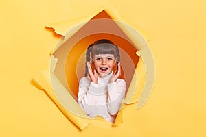 Horizontal shot of excited little girl with braids wearing casual shirt looking through torn hole in yellow paper, screaming with