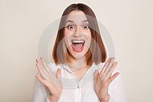 Horizontal shot of excited shocked woman wearing shirt posing isolated over white background, sees something amazed, raised arms,