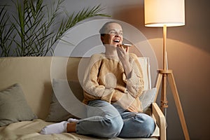 Horizontal shot of extremely happy young adult woman wearing casual clothing sitting on sofa and using smart phone, laughing