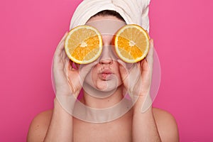 Horizontal shot of funny cheerful young woman holding two halves of orange against her eyes, having white towel on her head,