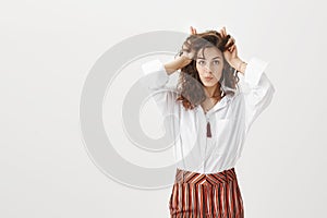 Horizontal shot of funny and cute feminine woman with curly hair, folding lips and gazing carefree at camera, holding