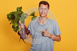 Horizontal shot of handsome model standing isolated over yellow background in gray t shirt, holding raw vegetable in hand, smiling