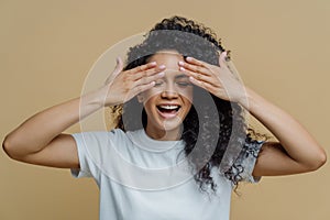Horizontal shot of happy dark skinned woman covers eyes and smiles, has fun and hides face, has curly hair, dressed in casual