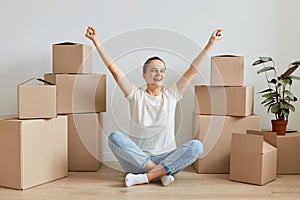 Horizontal shot of happy excited woman wearing white shirt and jeans posing at new apartment with carton boxes, sitting on floor