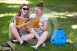 Horizontal shot of happy female studens feels relaxed while sit crossed legs on green grass, enjoy spring weather, have positive e