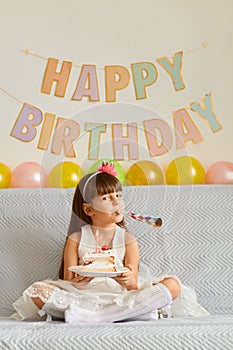 Horizontal shot of happy little girl wearing white dress sitting on sofa with birthday cake and blowing party horn, rejoicing her