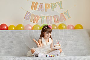 Horizontal shot of happy smiling little girl wearing white dress sitting on sofa with birthday cake and burning candle, rejoicing
