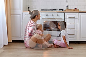 Horizontal shot of happy woman with bun hairstyle wearing striped shirt sitting on floor in kitchen with her kids, mother and her