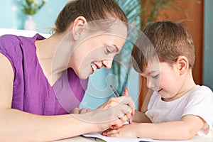 Horizontal shot of happy young smiling mother being glad to spend free time with her small son, draw something with great interest