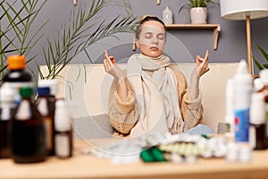Horizontal shot of ill Caucasian young adult woman sitting on cough in home interior among pills, practicing yoga, calming down