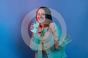 Horizontal shot of joyful adorable Caucasian young girl wearing casual jacket standing isolated over blue neon light background,