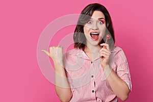 Horizontal shot of joyful attractive woman with makeup, holds lollipop, keeps mouth widely opened, wears stylish outfit, points