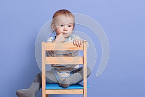 Horizontal shot of little boy sitting on stool, isolated on blue studio background, looks thoughtfuly aside, keeps finger near lip