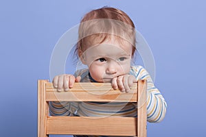 Horizontal shot of little child sitting in chair looking cartoons, funny toddler looks concentrated, posing isolated over blue