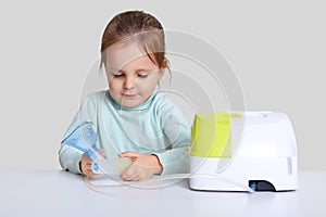 Horizontal shot of little girl sits at white desk with nebulous machine for health care, holds mask for making inhalation or