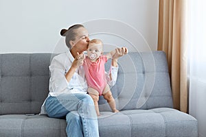 Horizontal shot of lovely woman with bun hairstyle wearing white shirt and jeans, young mother kissing her infant daughter while