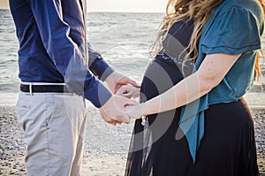 Horizontal shot of a man in blue shirt and a pregnant woman in black dress holding hands on beach