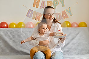 Horizontal shot of mother sitting on sofa with her toddler daughter and holding cake with candles, being ready blowing together,