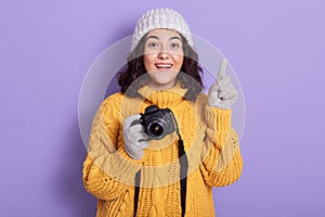 Horizontal shot of photographer girl over lilac wall showing and pointing finger up, having great idea, looking directly at camera