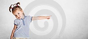 Horizontal shot of pretty lovely beautiful small kid wears striped t shirt, has funny two pony tails, pointing with her little han