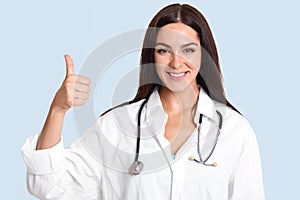 Horizontal shot of pretty talented female doctor with satisfied expression, raises thumb, shows like gesture, approves something,